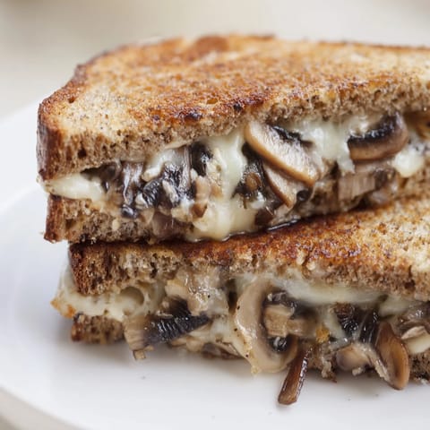 Two halves of a savory Mushroom Swiss Grilled Cheese are stacked on a plate, ready to be dipped into a bowl of warm tomato soup.