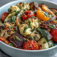 Brightly colored roasted vegetables, including red peppers and purple cauliflower, sit atop fluffy brown rice in a vegan Rainbow Roasted Vegetable Bowl. A vibrant green herb sauce is drizzled over the top.